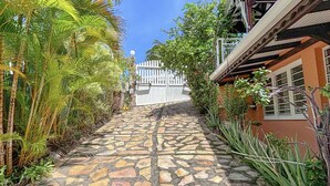 Un chemin de pierre mène à un portail blanc, bordé de plantes tropicales et d’un bâtiment à la façade rouge.