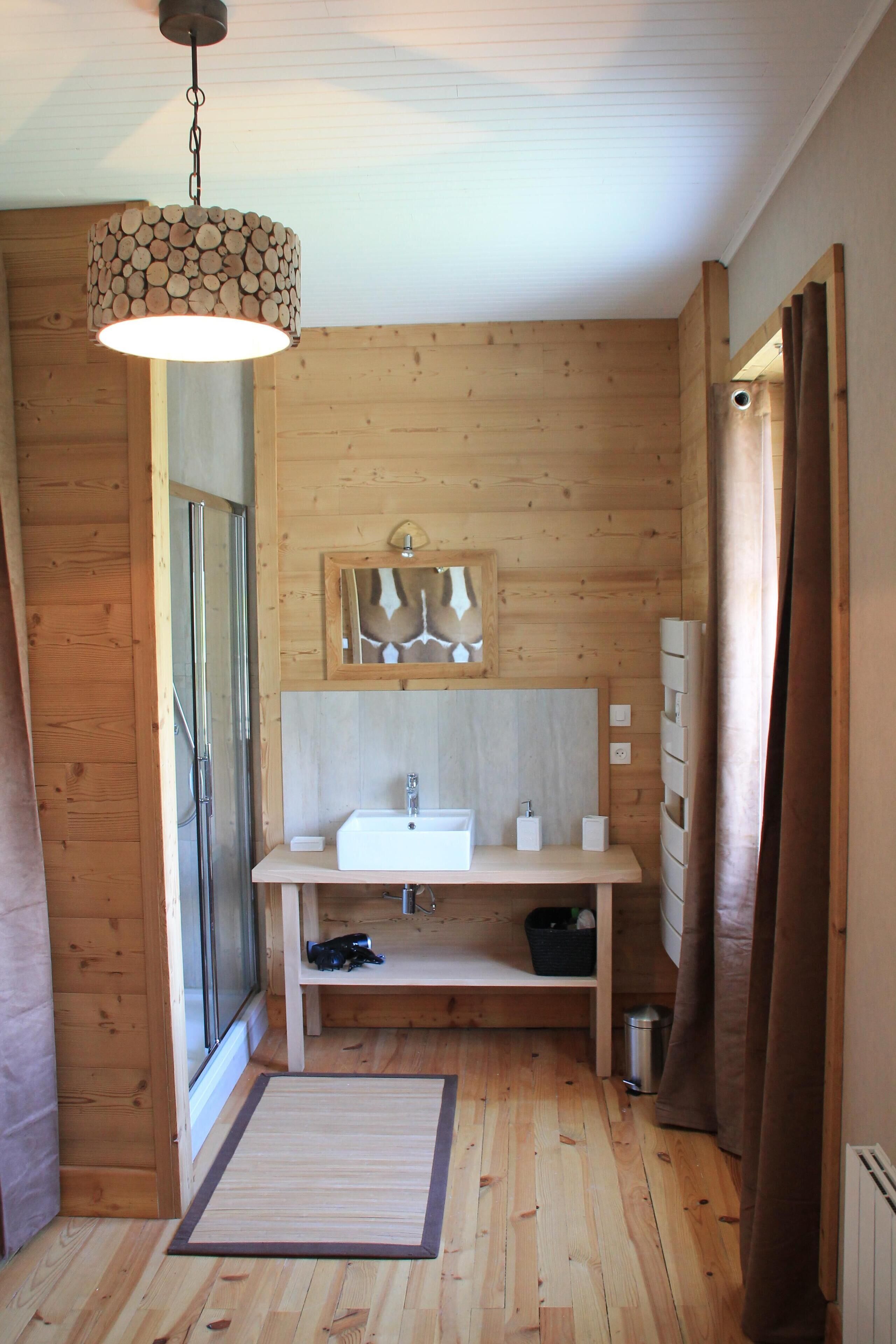 Une salle de bain rustique, avec des murs et un plafond en bois, un luminaire suspendu, un lavabo, un miroir et une douche.
