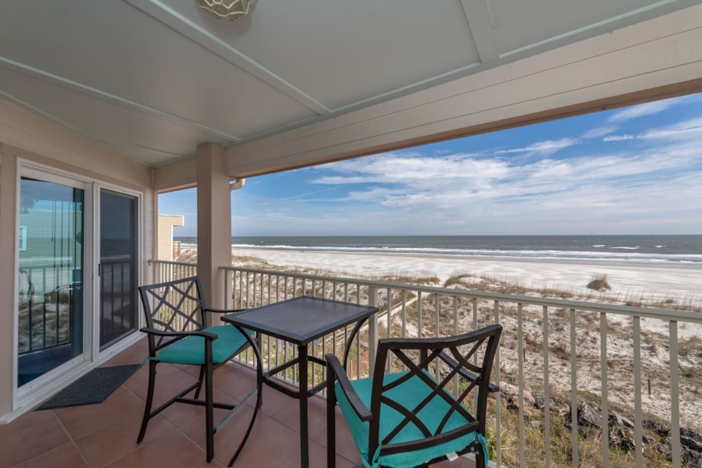 A balcony with a table and chairs, overlooking a beach with sand dunes and the ocean.