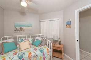 A bedroom with a bed, a nightstand, a lamp, a beach-themed wall art, a window with blinds, and a door.