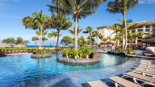 A resort pool area with palm trees, lounge chairs, and a view of the ocean.