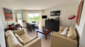 A living room with two beige sofas, a wooden coffee table, a dining table with chairs, a TV mounted on the wall, and a balcony with a view. - Renovated apartment in Valescure, with pool and golf view, near the sea  (Saint-Raphaël)