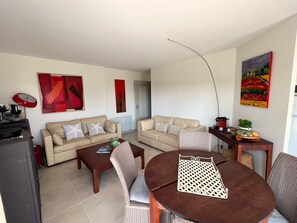 A living room with two sofas, a coffee table, and a dining table with chairs. There are abstract paintings on the walls and a plant on the side table.