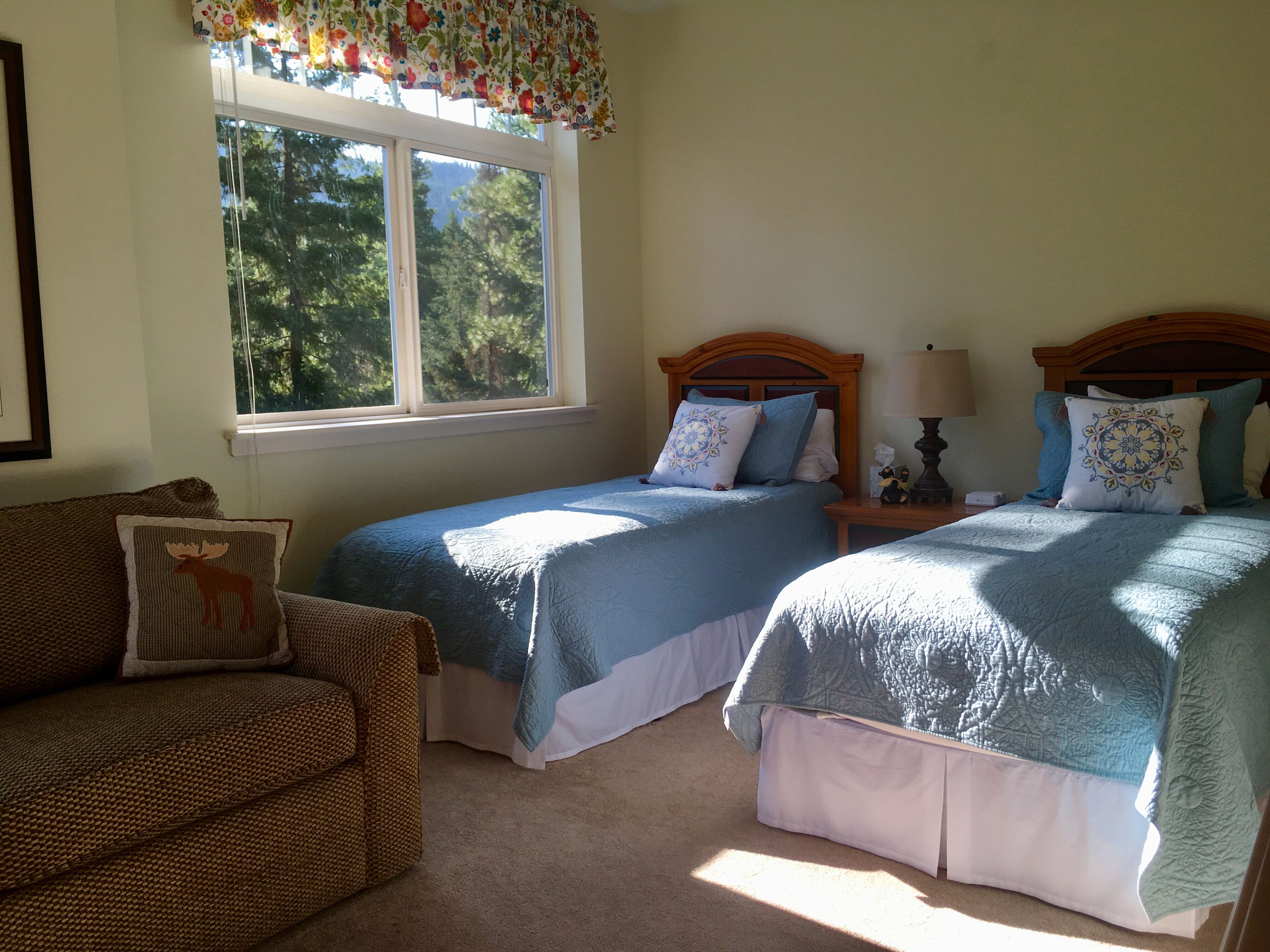 A bedroom with two beds, a window with a view of trees, a wooden headboard, and a small table with a lamp.