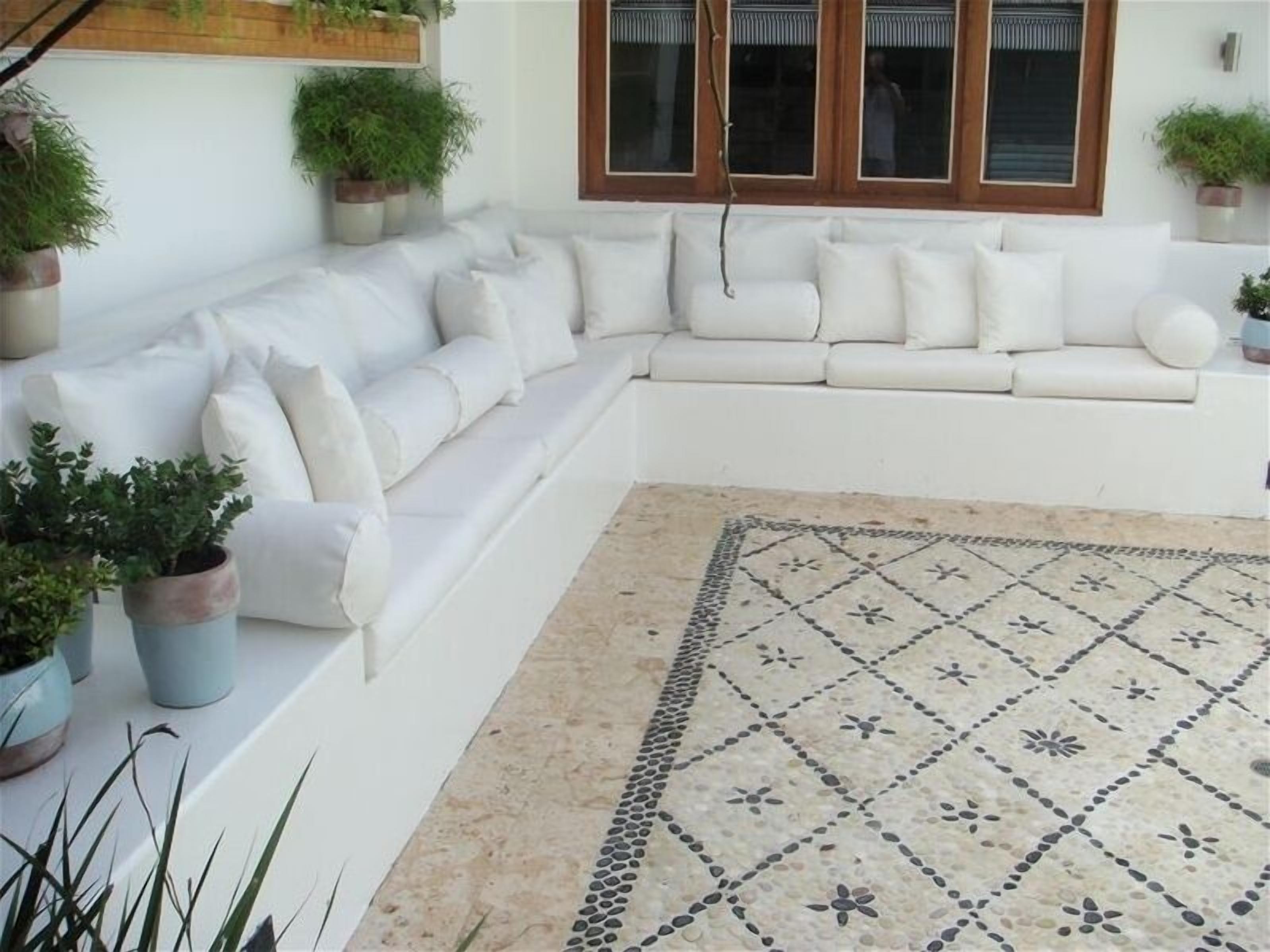 Een witte bank met kussens, potplanten en een vloerkleed met patroon in een kamer met een raam en wandplanten.
