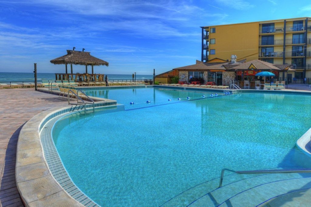 Une piscine d’un bleu éclatant, dotée d’une terrasse en bois et d’un parasol en chaume, le tout entouré d’un bâtiment jaune agrémenté de balcons.