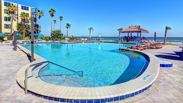 A swimming pool with a curved edge, surrounded by lounge chairs and palm trees, with a beach and ocean in the background. - Oceanfront - Poolside - Steps to Beach & Tiki Bar (Daytona Beach Shores)