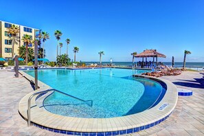 Ein Pool mit geschwungener Kante, umgeben von Liegestühlen und Palmen, mit Strand und Meer im Hintergrund.