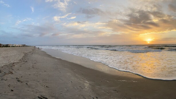 A beach with a wide sandy path leading to the water, buildings in the background, and a sunset over the horizon. - Lovely Ocean Views! (Cocoa Beach)