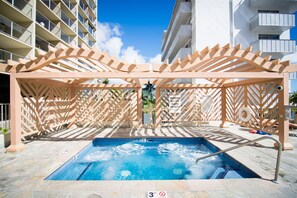 Área de piscina com hidromassagem, cercada por uma pérgola de madeira, e uma placa indicando ausência de salva-vidas.