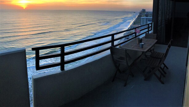 A balcony with a view of the ocean at sunset, wooden furniture, and a railing. - Luxury Ocean Front 2br/2ba Horizons Condo17th Floor - Awesome views (Daytona Beach)