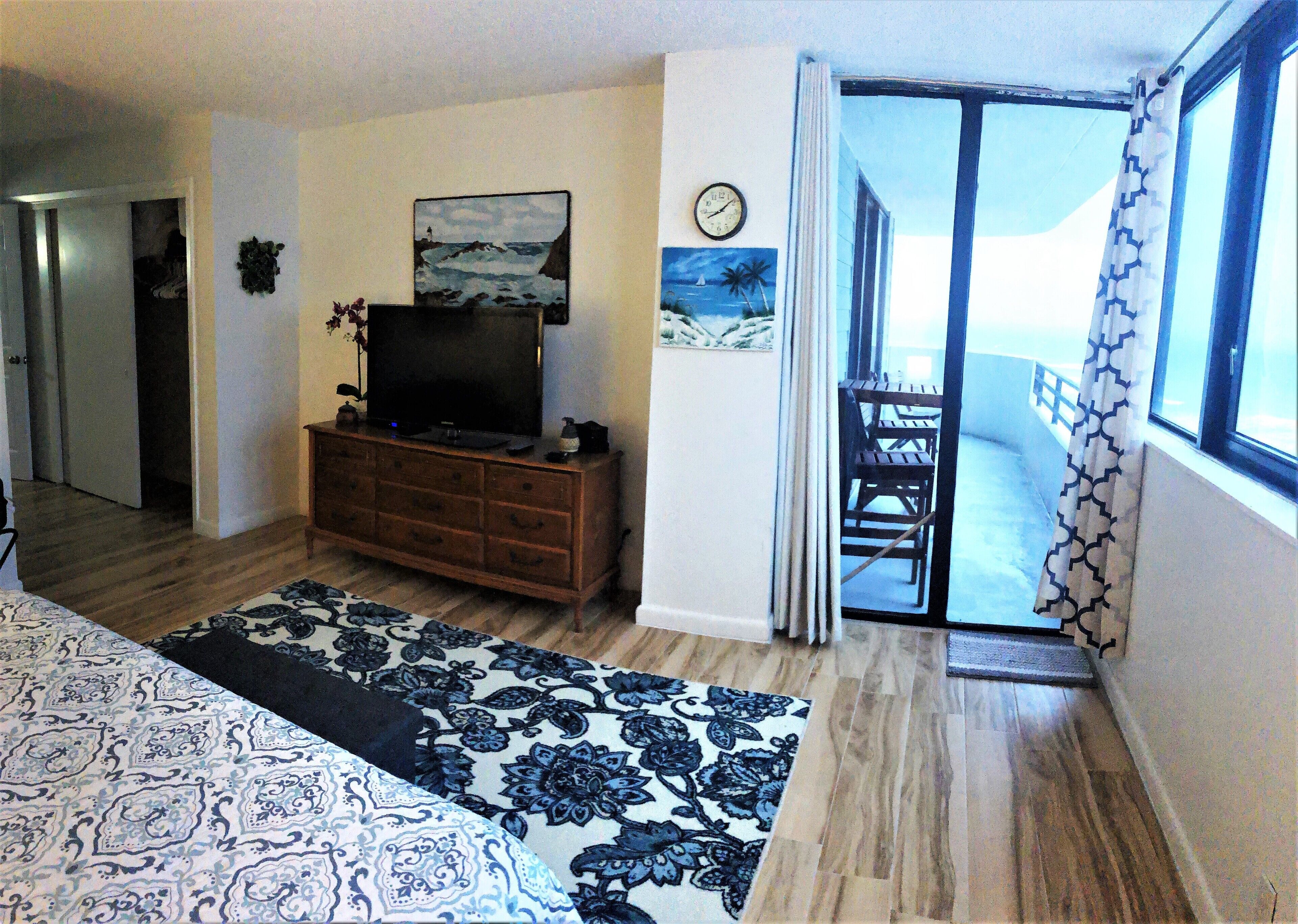 Un dormitorio con cama, cómoda, televisor, reloj y balcón con vista al mar.
