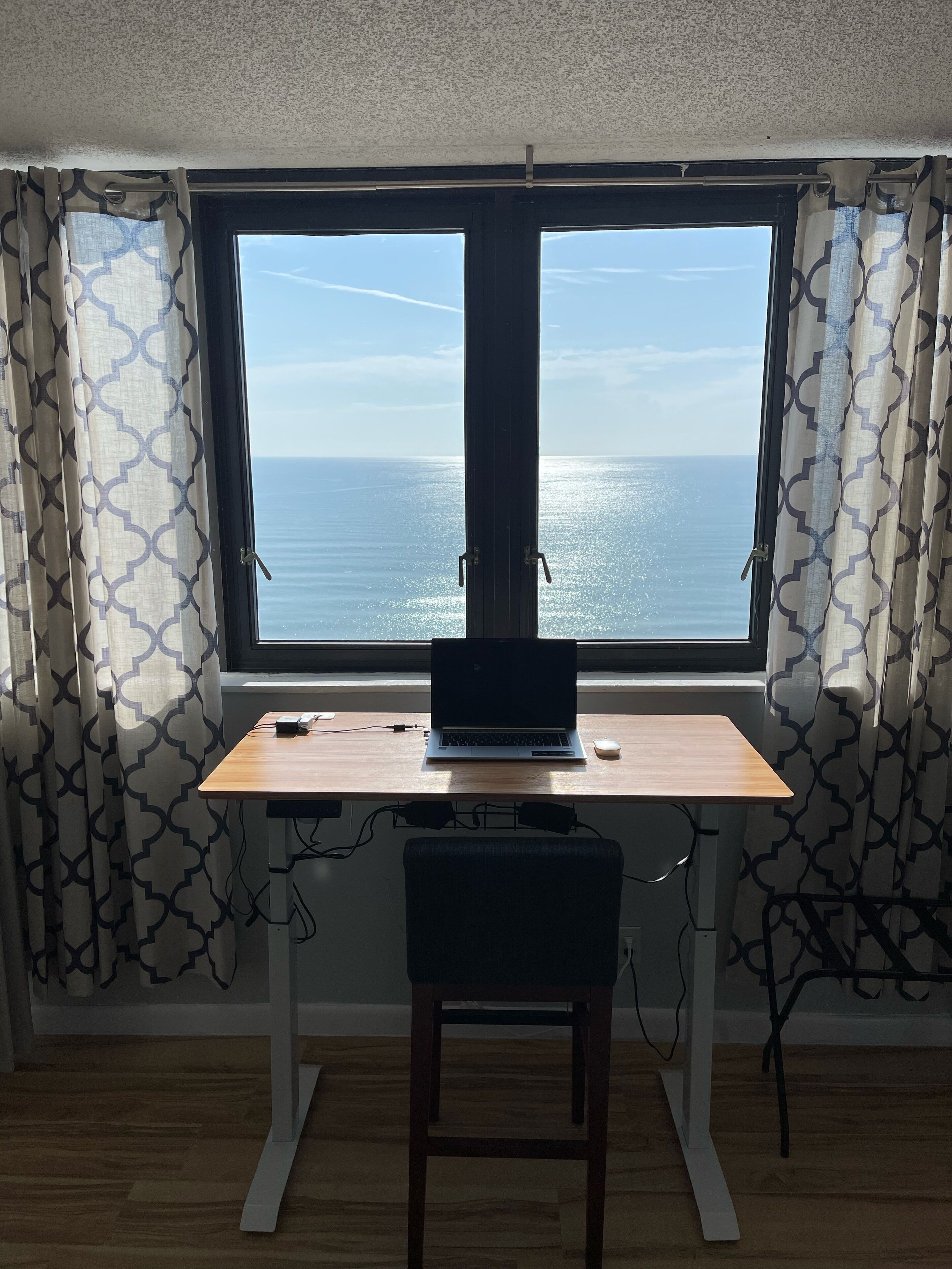 Una habitación con escritorio y silla, una computadora portátil y vista al mar a través de un ventanal grande.
