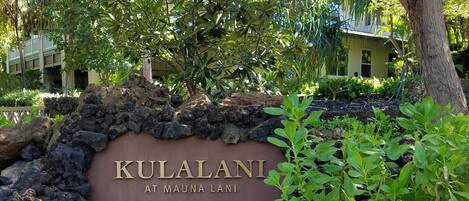 Um letreiro com 'KULALANI AT MAUNA LANI' em um ambiente natural, com vegetação e uma árvore.