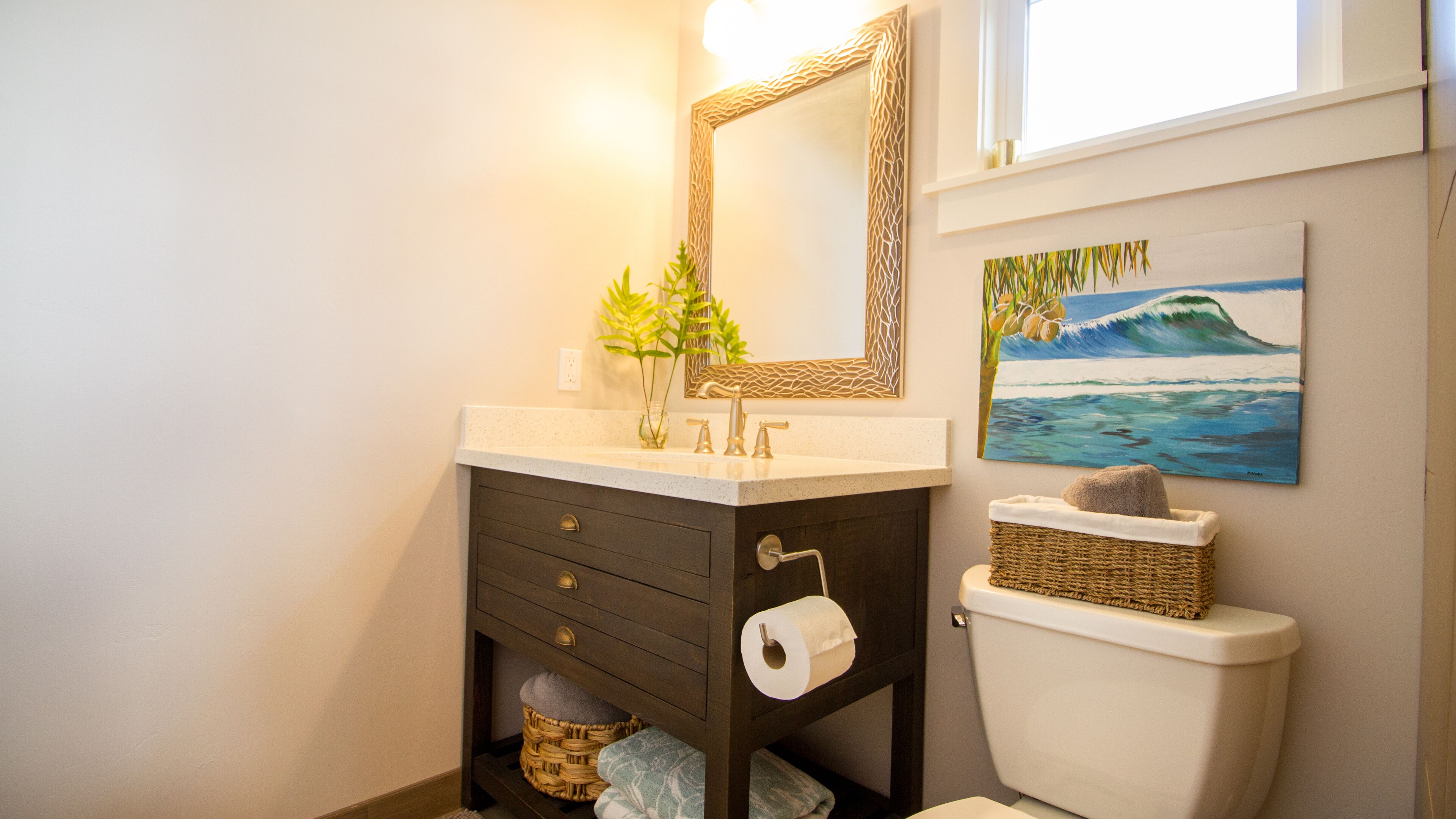 Une salle de bain avec un lavabo blanc, un meuble-lavabo en bois foncé, un miroir, des toilettes, un panier et un tableau représentant une vague.