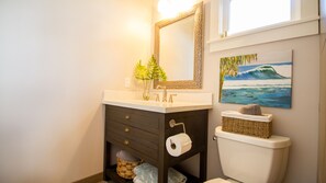 A bathroom with a white sink, dark wooden vanity, a mirror, a toilet, a basket, and a painting of a wave.