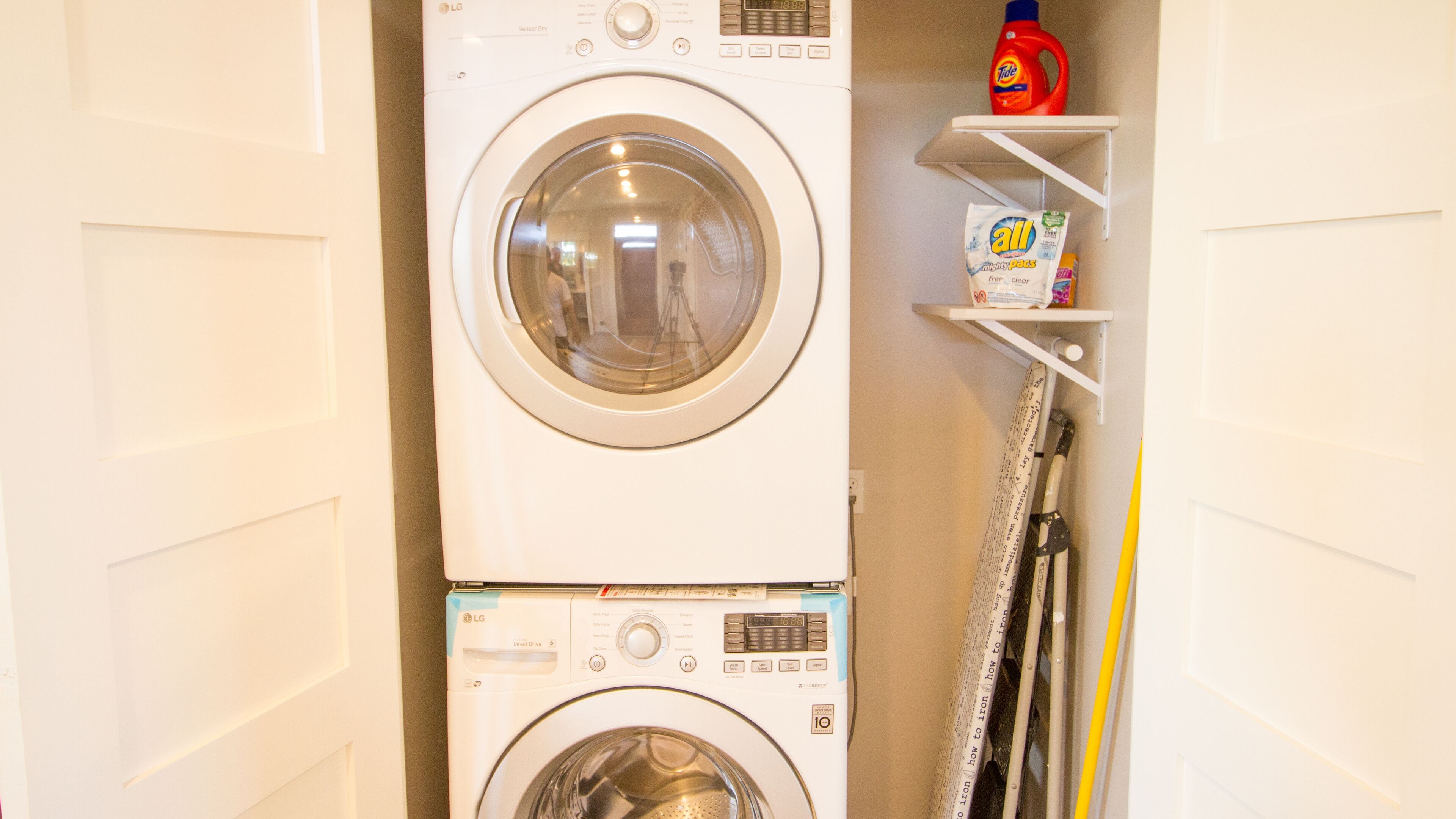 Een gestapelde wasmachine en droger, met daarboven een plank waarop een fles Tide wasmiddel en een doos All wasmiddel staan.
