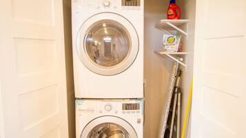 Een gestapelde wasmachine en droger, met daarboven een plank waarop een fles Tide wasmiddel en een doos All wasmiddel staan.