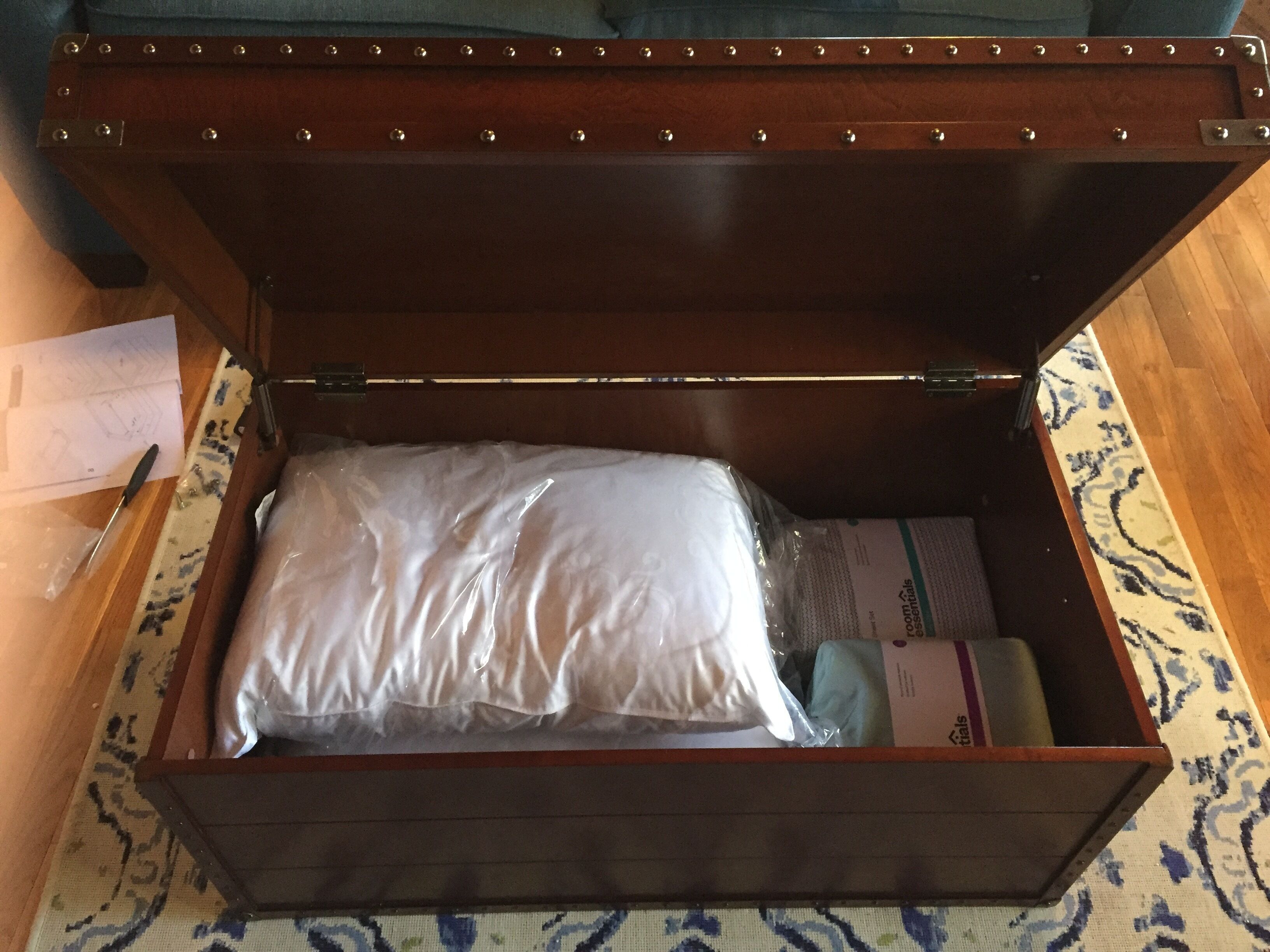 A wooden chest with a hinged lid, containing a white pillow, a rolled-up towel, and a box of toiletries.