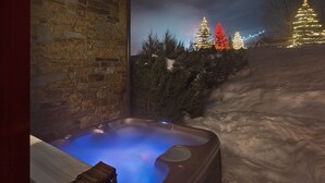 Een bubbelbad met een bakstenen muur op de achtergrond, omringd door sneeuw en verlichte kerstbomen.