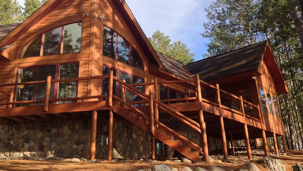 A wooden cabin with a stone foundation, large windows, and a wooden staircase leading to a covered porch.