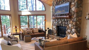 A cozy living room with a stone fireplace, comfortable seating, and a large window overlooking a forest.
