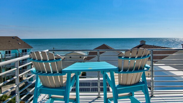 Un balcon avec deux chaises et une table, donnant sur une plage et l’océan.