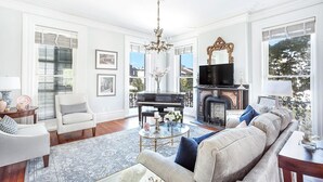 A spacious living room with a grand piano, a fireplace, and a large window offering a view of the outdoors. - Grand Manor on Broughton Street w/ Parking, Courtyard by Lucky Savannah (Savannah)
