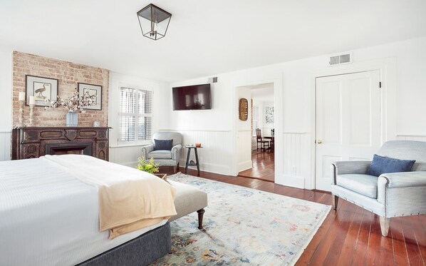 A bedroom with a large bed, a fireplace, a TV, and a dining area in the background. - Grand Manor on Broughton Street w/ Parking, Courtyard by Lucky Savannah (Savannah)
