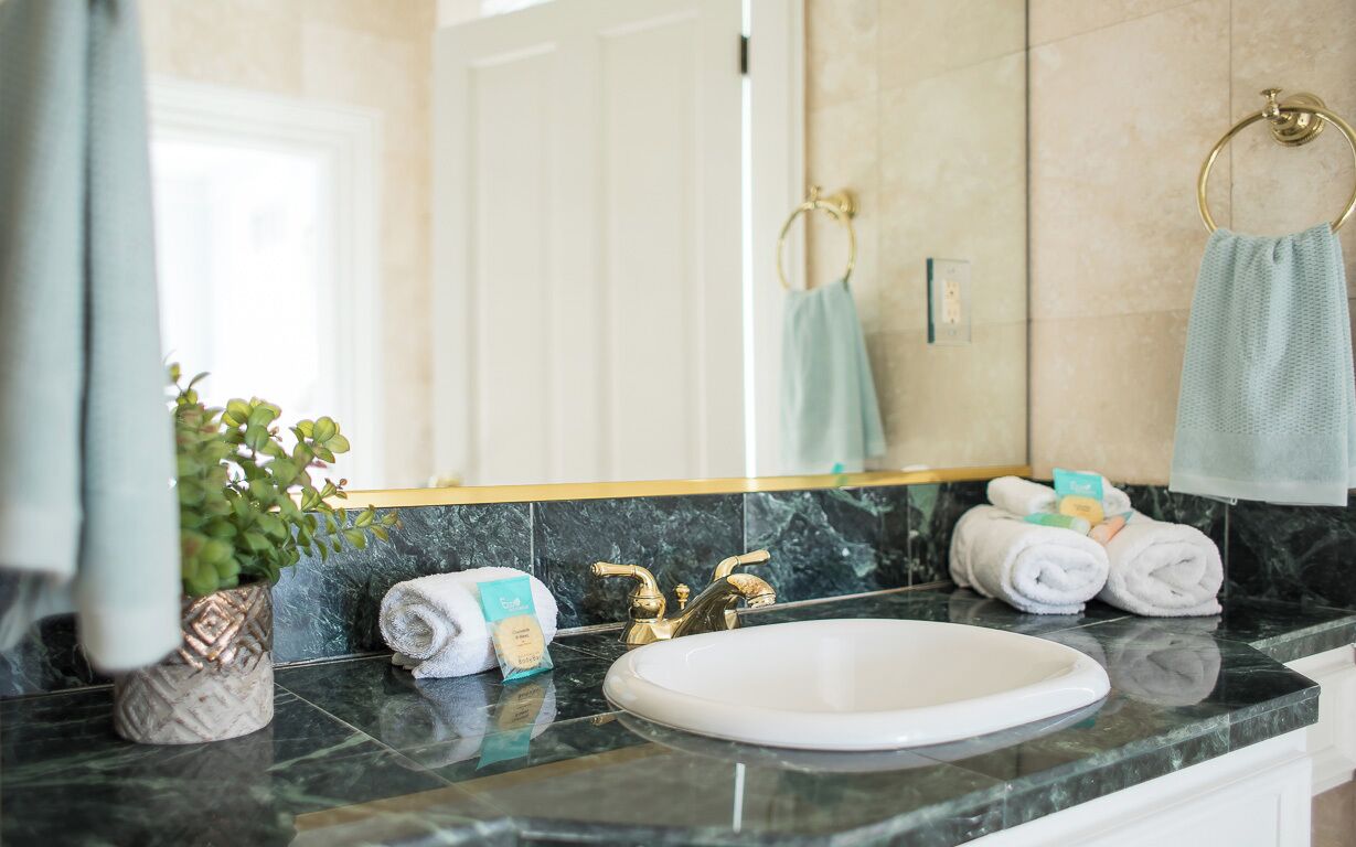 Ein Badezimmer mit weißem Waschbecken, goldenen Wasserhähnen, einer Marmorarbeitsplatte, einer Topfpflanze und Handtüchern.