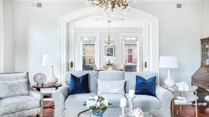 A living room with a light grey sofa, blue cushions, a glass coffee table, and a chandelier. - Grand Manor on Broughton Street w/ Parking, Courtyard by Lucky Savannah (Savannah)