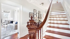 A grand staircase with a wooden handrail and white balusters, leading to a landing with framed artwork and a chandelier. - Grand Manor on Broughton Street w/ Parking, Courtyard by Lucky Savannah (Savannah)