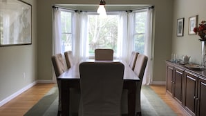 A dining room with a long table and chairs, a chandelier, and large windows with curtains. - Beautiful North Fork 3-Bedroom Home Steps Away from a Private Sandy Bay (Southold)