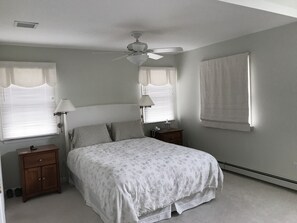 Une chambre à coucher comprenant un lit, deux tables de chevet, un ventilateur de plafond et une fenêtre avec des stores.