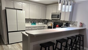 A kitchen with white cabinets, a stainless steel refrigerator, a microwave, a kettle, and a bar with stools. - Spacious 3bd 3ba, 1 min walk to private beach with 2 master king bed (Miramar Beach)