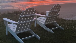 Dos sillas Adirondack en un área cubierta de césped con vistas a una playa al atardecer.