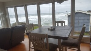 Un comedor con mesa y sillas, ventanas amplias con vista al océano y un cobertizo azul al fondo.