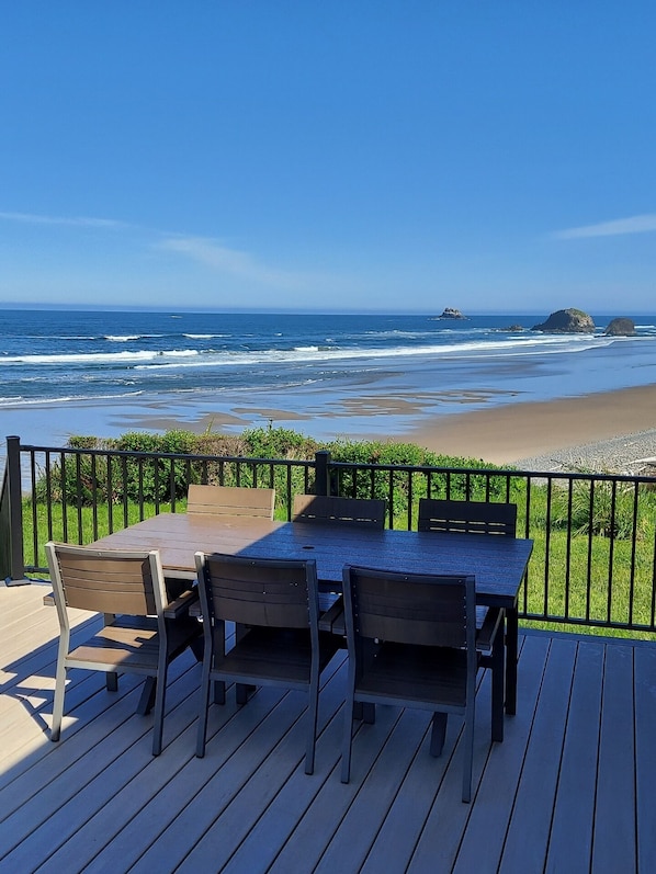 A wooden deck with a table and chairs overlooking a beach with waves and rocks.