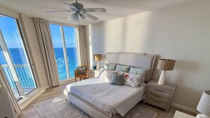 A bedroom with a large bed, a ceiling fan, a nightstand, a lamp, and a view of the ocean through large windows.