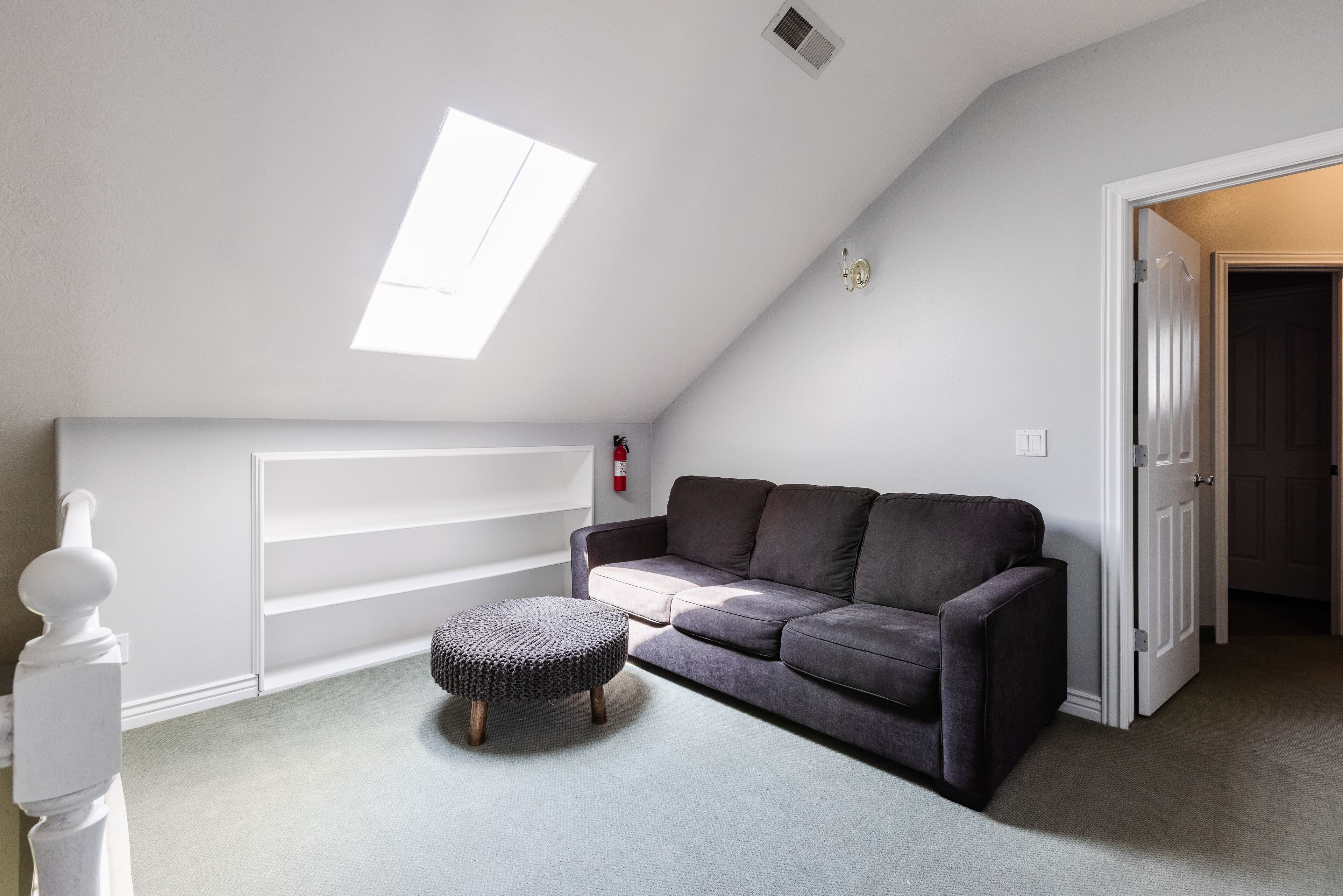 A room with a sloped ceiling, a grey sofa, a round ottoman, and a white wall with a shelf.