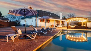 Une piscine avec des chaises longues et des parasols, un patio couvert et une maison en arrière-plan.