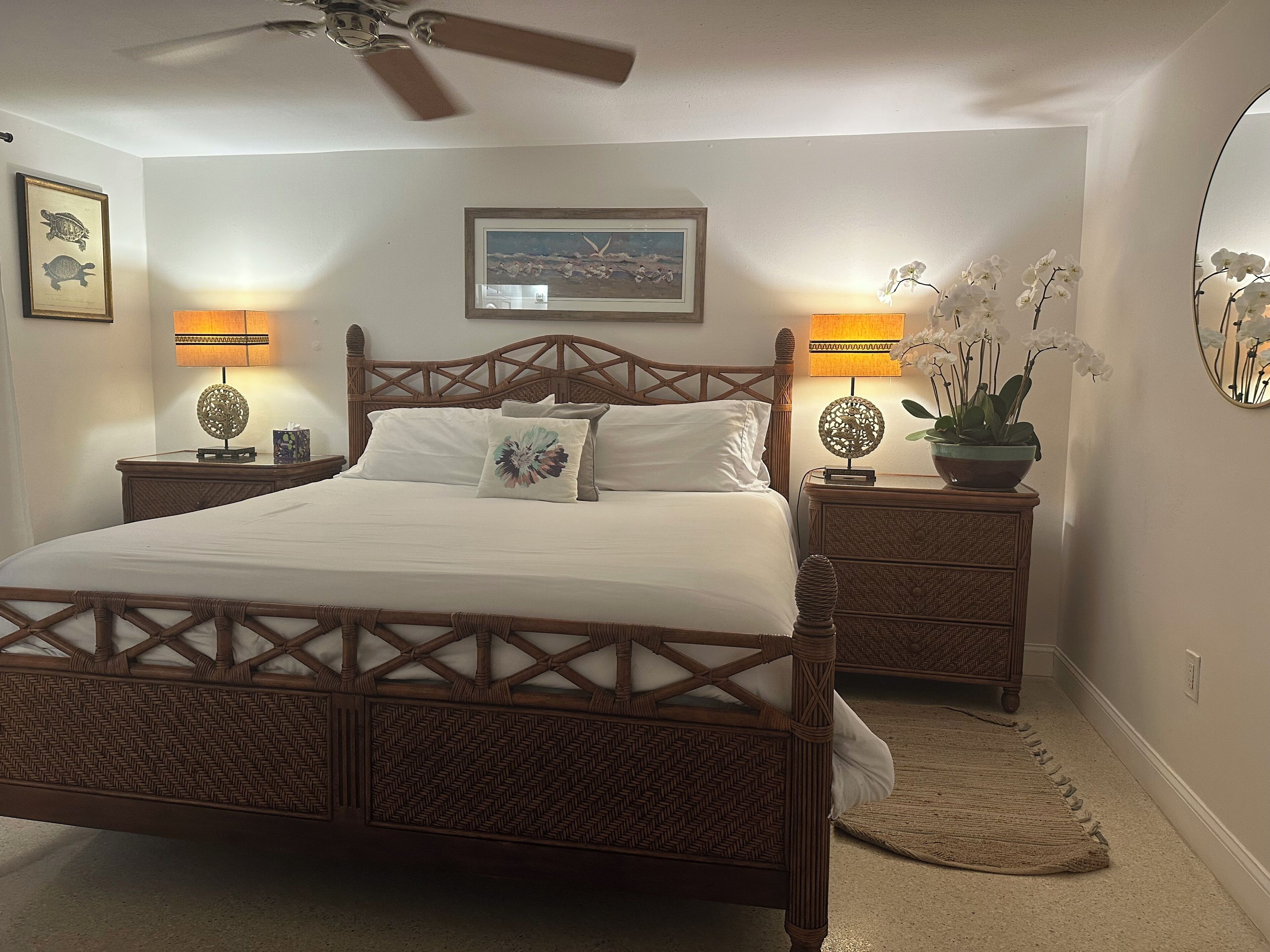 A bedroom with a wooden bed, two bedside tables with lamps, a ceiling fan, a framed picture, and a potted orchid.