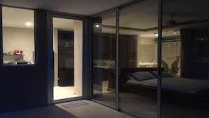 Un dormitorio moderno con una puerta de cristal que da a un baño, una cama con almohadas y una pequeña zona de kitchenette visible a través de la puerta.