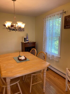 A dining area with a wooden table, chairs, a chandelier, a window with lace curtains, and a sign on the wall.