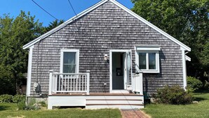 A single-story house with a front porch, a small attached garage, and a brick pathway leading to the entrance. - Beautiful 2 Bedroom Cottage Just Short Stroll To Town Neck Beach (Sandwich)