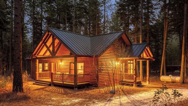 A cabin with a wooden exterior, a porch, and a metal roof, surrounded by a forest.