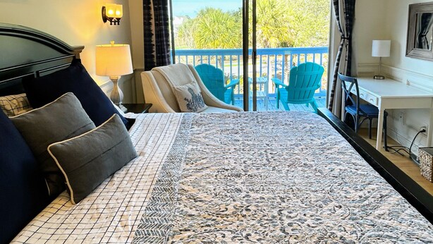 A hotel room with a bed, a desk, a chair, a balcony with chairs, and a view of trees.