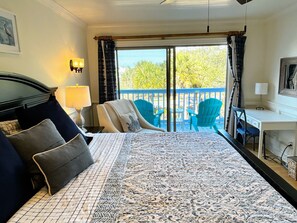 A hotel room with a bed, a desk, a chair, a balcony with chairs, and a view of trees.