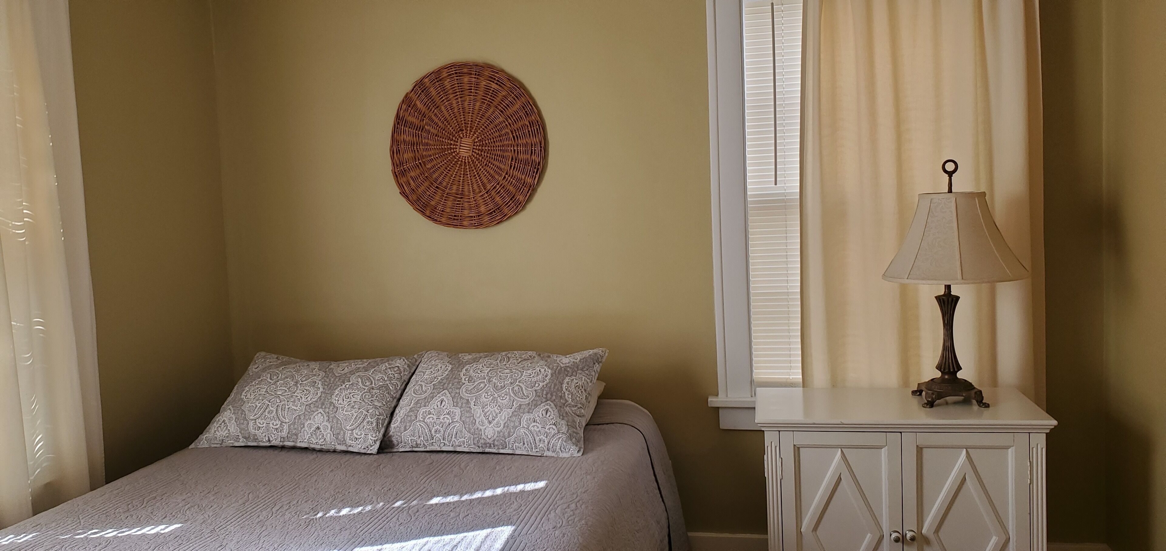 Une chambre à coucher avec un lit, deux coussins à motifs, une décoration murale, une table de chevet avec une lampe et une fenêtre avec des rideaux.
