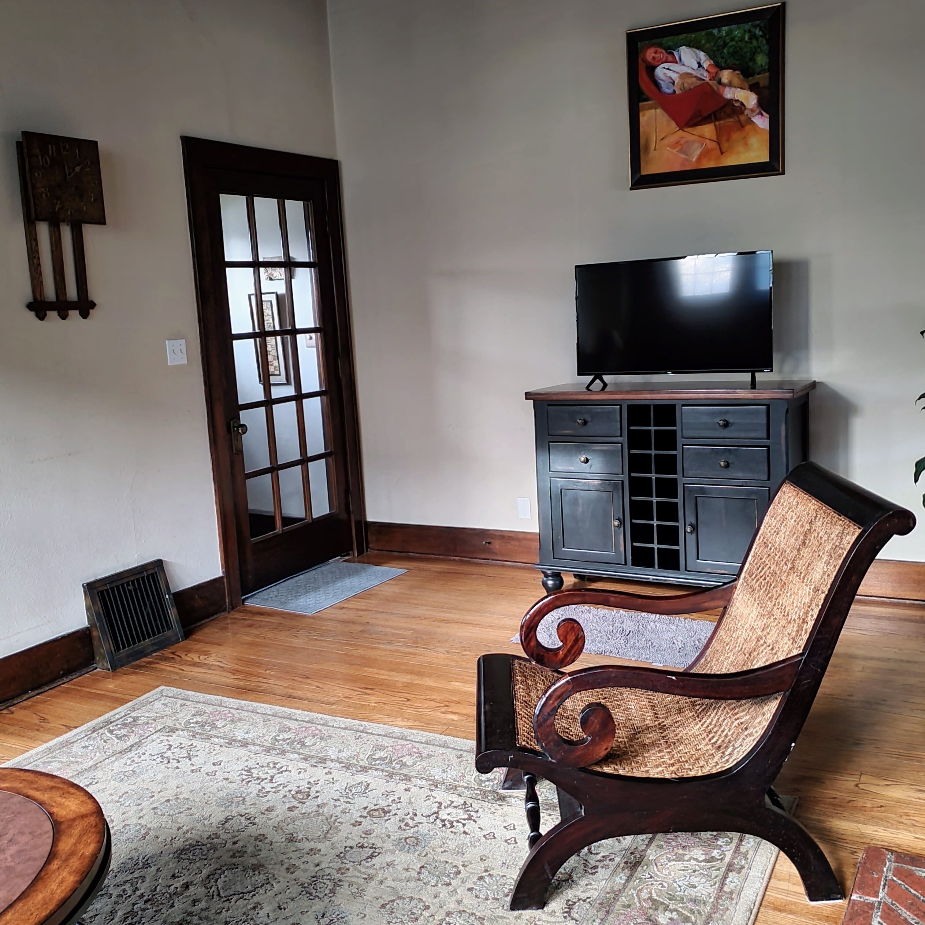 A living room with a wooden floor, a patterned rug, a dark wooden chair, a television on a cabinet, and a door with frosted glass panels.